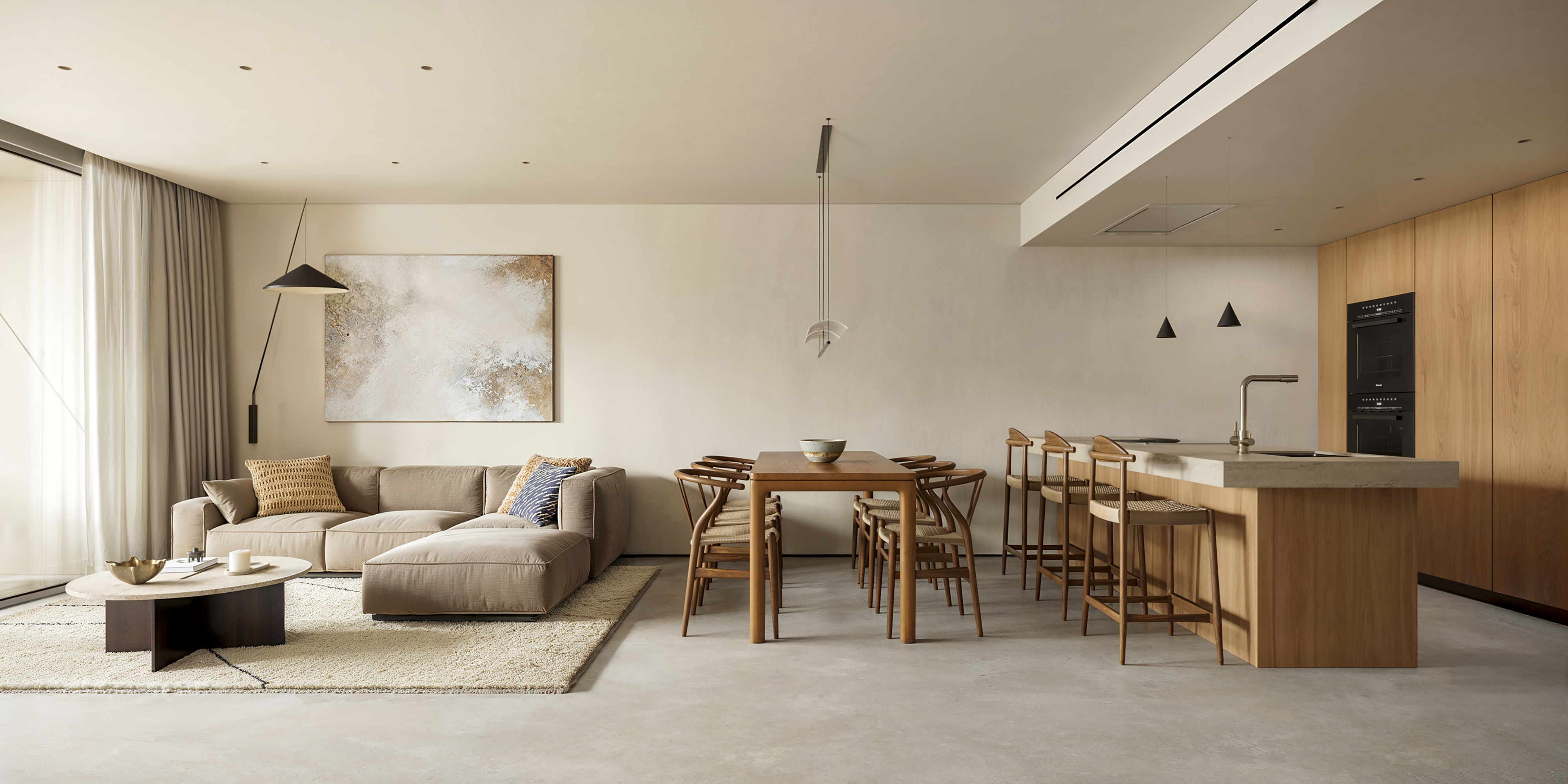 Espai interior obert i lluminós d'estil minimalista i tons neutres, que integra de forma contínua una sala d'estar amb un sofà cantoner, una taula de menjador de fusta i una cuina moderna amb illa central i tamborets.